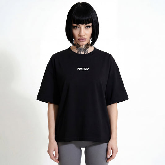 Front view of a model with tattoos and sharp makeup wearing the RAVEDRIP Core Matrix Boxy Tee in black, featuring a relaxed fit and minimalist chest logo in an industrial studio setting.