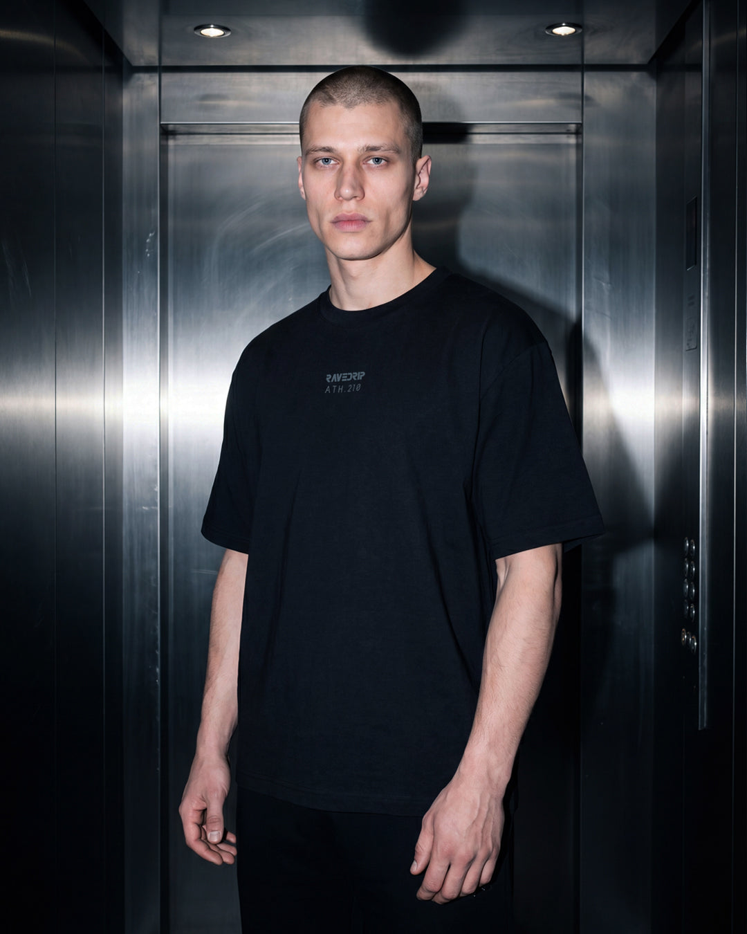 A man with a buzz cut stands inside a metallic elevator, wearing a black heavyweight t-shirt with a small white printed logo RAVEDRIP
