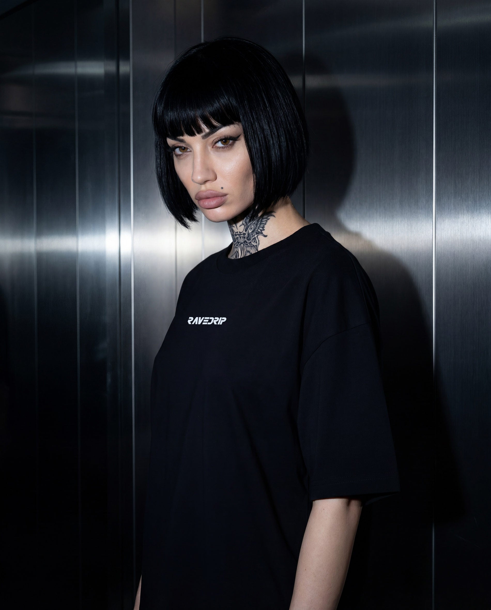 A model with a black bob haircut and neck tattoos wearing an oversized black t-shirt with a small white RAVEDRIP logo on the chest, standing against a metallic dark gray wall.