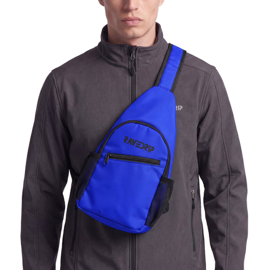 Close-up cropped studio shot of a RAVEDRIP royal blue canvas sling chest bag with a vertical teardrop design. The bag features a black zipper, mesh side pockets, and a black logo, worn front-facing over a technical charcoal grey softshell jacket. Premium e-commerce techwear product photography.