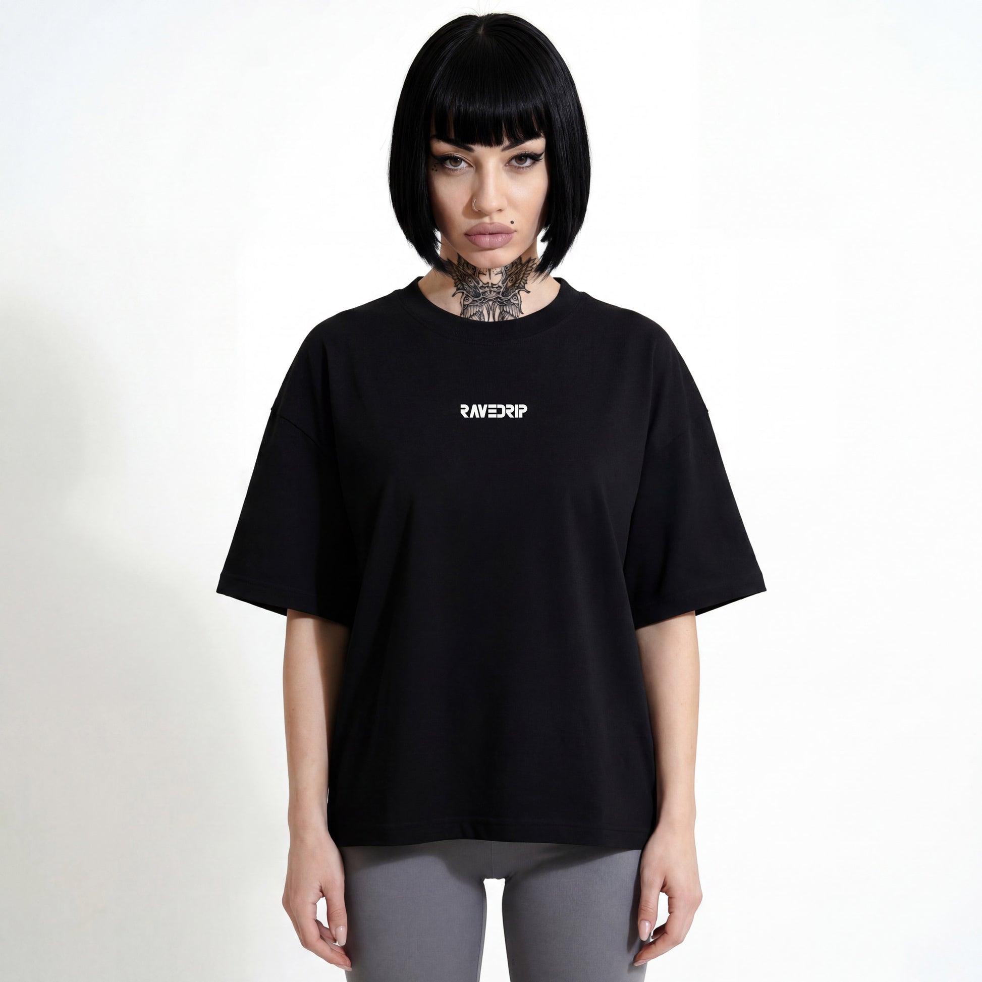 Front view of a model with tattoos and sharp makeup wearing the RAVEDRIP Core Matrix Boxy Tee in black, featuring a relaxed fit and minimalist chest logo in an industrial studio setting.