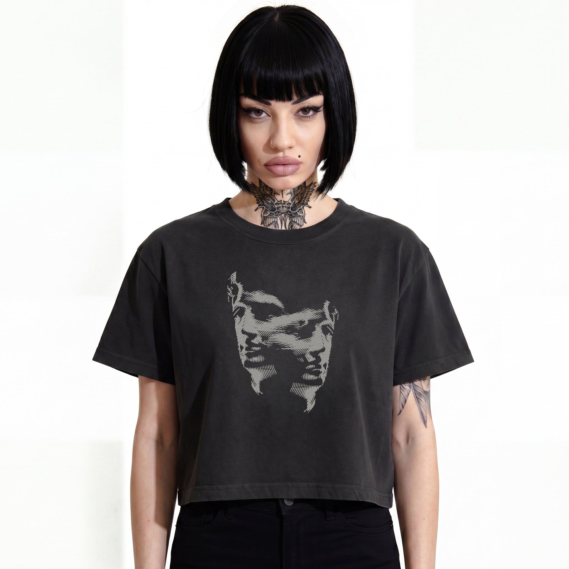 RAVEDRIP Duality heavyweight boxy cropped tee in faded black, premium wide square fit women's streetwear t-shirt with a grey engraved Janus-style double face duality graphic print on the chest, worn by alternative female model with a black bob haircut and neck tattoo, professional e-commerce studio catalog photography on a white background.
