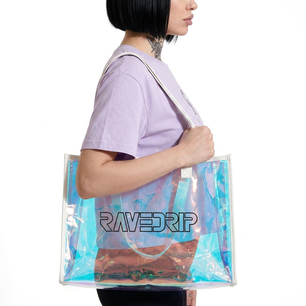 Side profile view of a model wearing a RAVEDRIP relaxed-fit lilac t-shirt, carrying a large transparent holographic turquoise vinyl tote bag on her shoulder. The glossy iridescent PVC material reflects blue and pink light, showcasing the RAVEDRIP black outline logo. Premium streetwear product photography.
