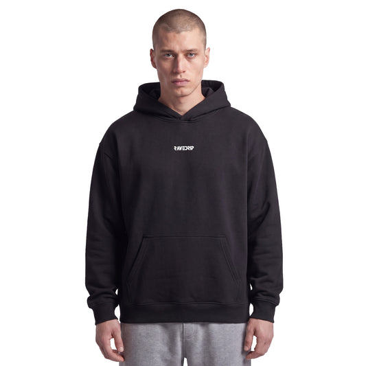 Studio lookbook portrait of male model wearing the RAVEDRIP Core Relaxed heavyweight hoodie in Black. The 9.4oz AS Colour fleece hoodie features a drawstring-less double-layer hood, dropped shoulders, and a small minimalist white logo printed on the high chest, styled for an edgy, iconic streetwear look.